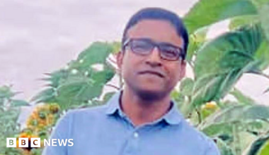 A man in his 40s with short, black hair wearing a blue polo shirt and glasses standing in front of plants