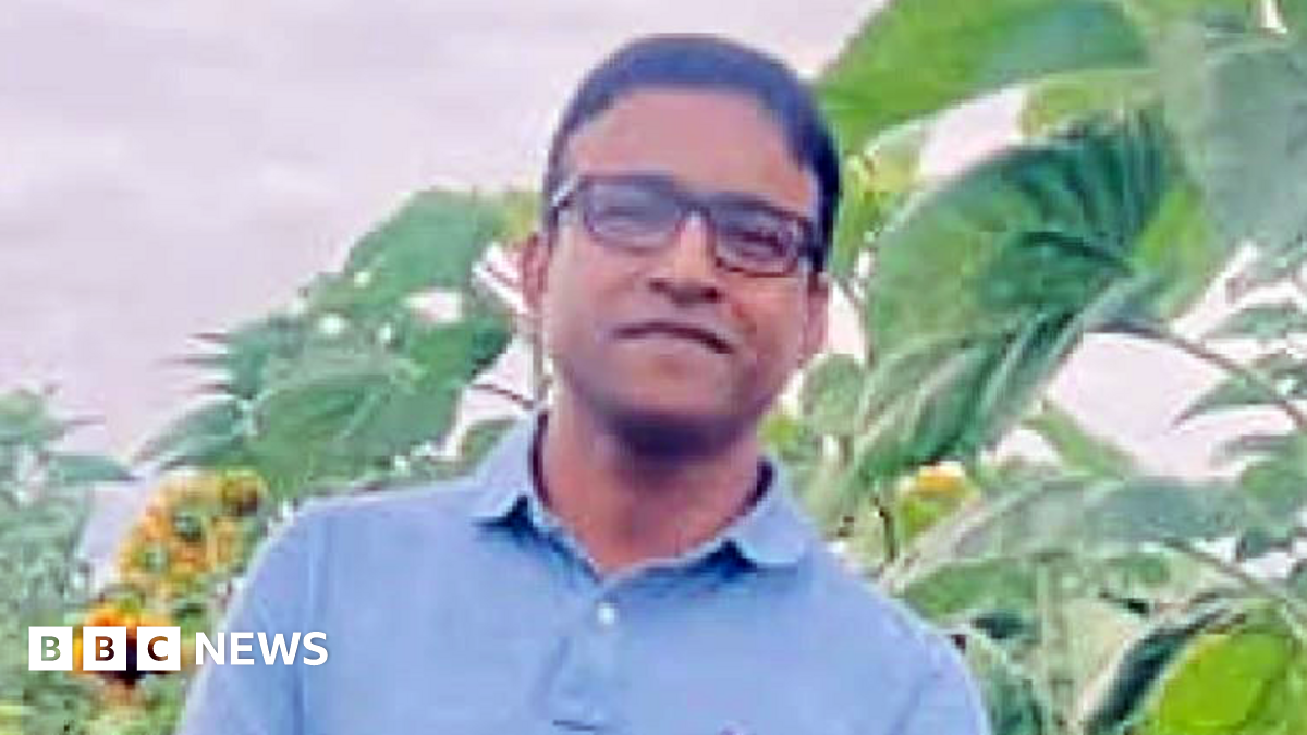 A man in his 40s with short, black hair wearing a blue polo shirt and glasses standing in front of plants