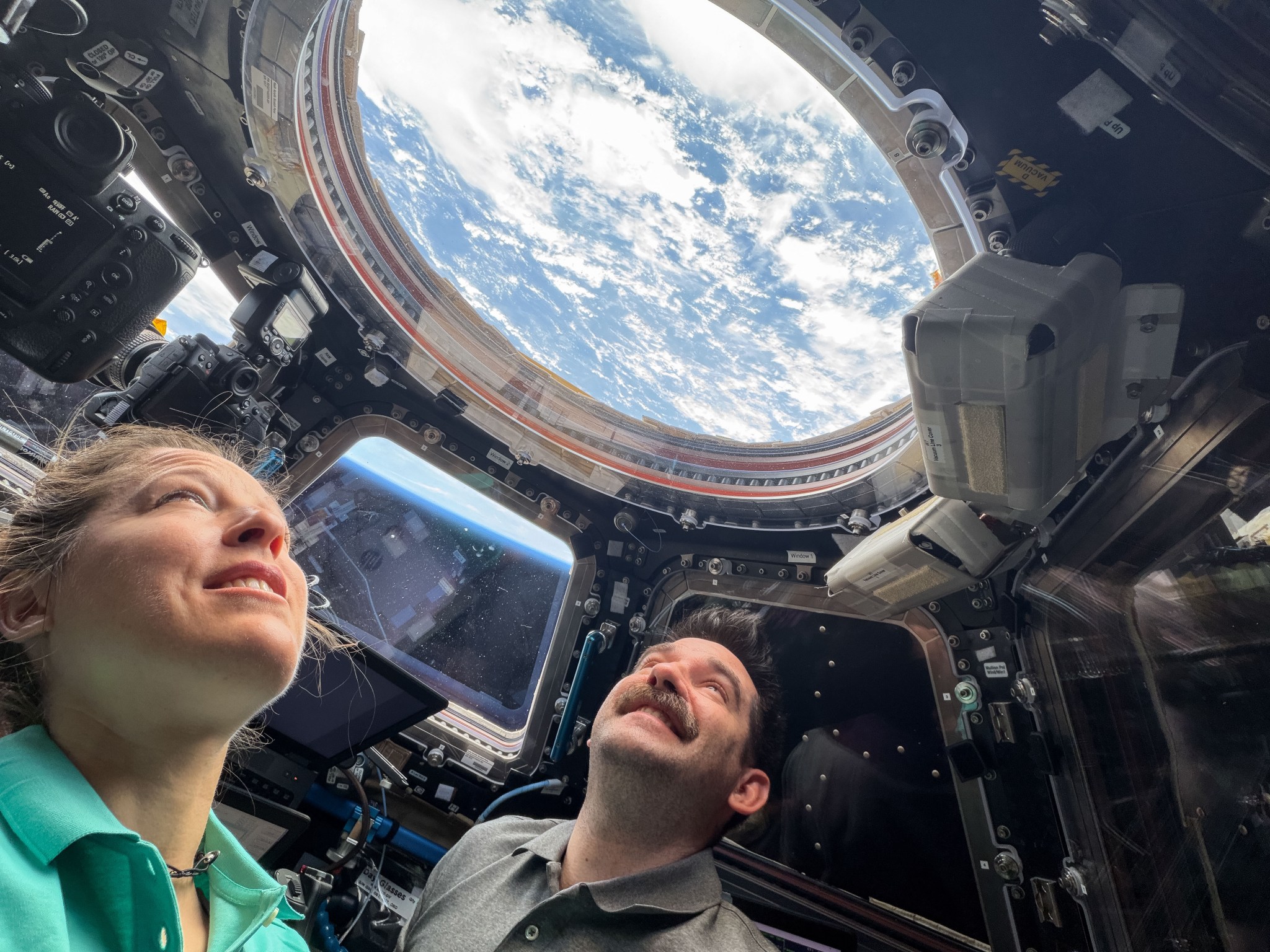 Two astronauts look up through a round window in the International Space Station's cupola. Earth's blue water and white clouds are visible. In a trapezoid-shaped window between the two astronauts, we can see the blue haze of Earth's atmosphere.