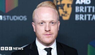 Owain Rhys Davies is smiling at the camera, he has red hair and beard. He wears a white shirt and black jacket.
