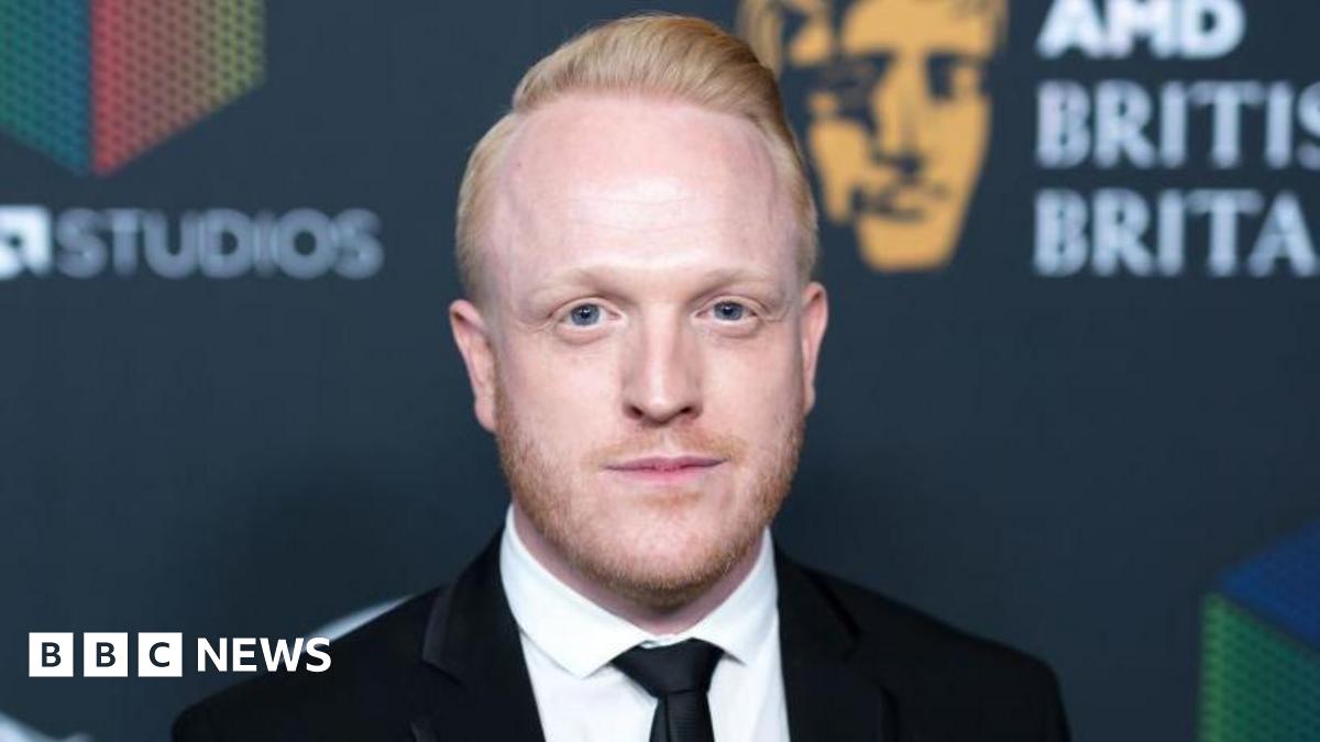 Owain Rhys Davies is smiling at the camera, he has red hair and beard. He wears a white shirt and black jacket.