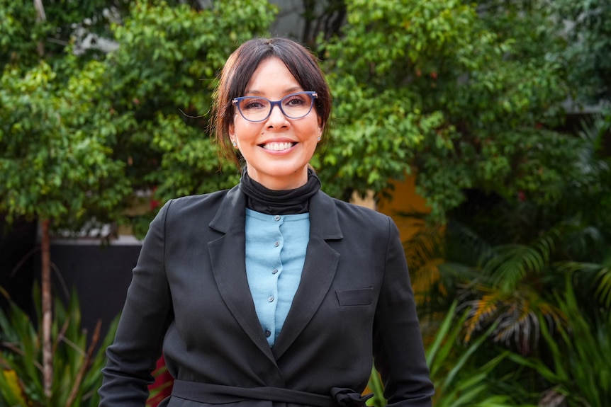Tatum Maybir in a black suit, blue shirt, glasses, smiling in front of greenery.