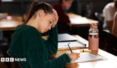A teenage girl writing on paper while sitting at a desk during exam in classroom. She has one hand on her head and has a concentrated expression. She wears a green jumper and has dark hair pulled back in a ponytail.