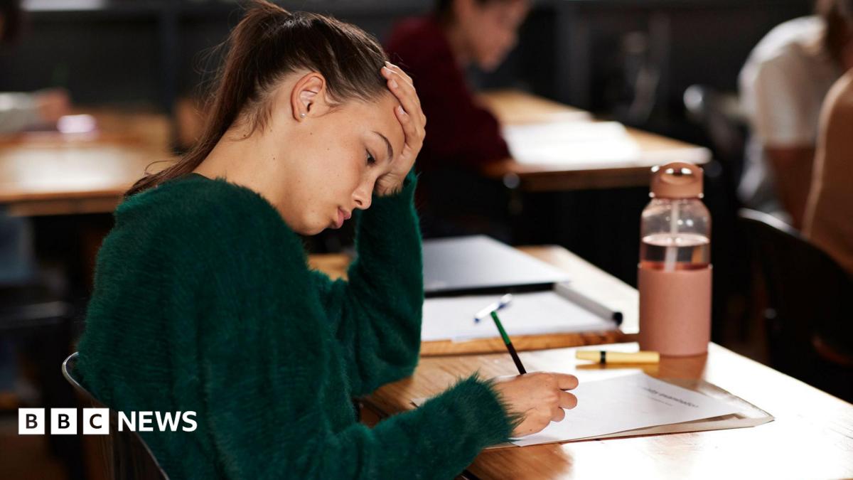 A teenage girl writing on paper while sitting at a desk during exam in classroom. She has one hand on her head and has a concentrated expression. She wears a green jumper and has dark hair pulled back in a ponytail.