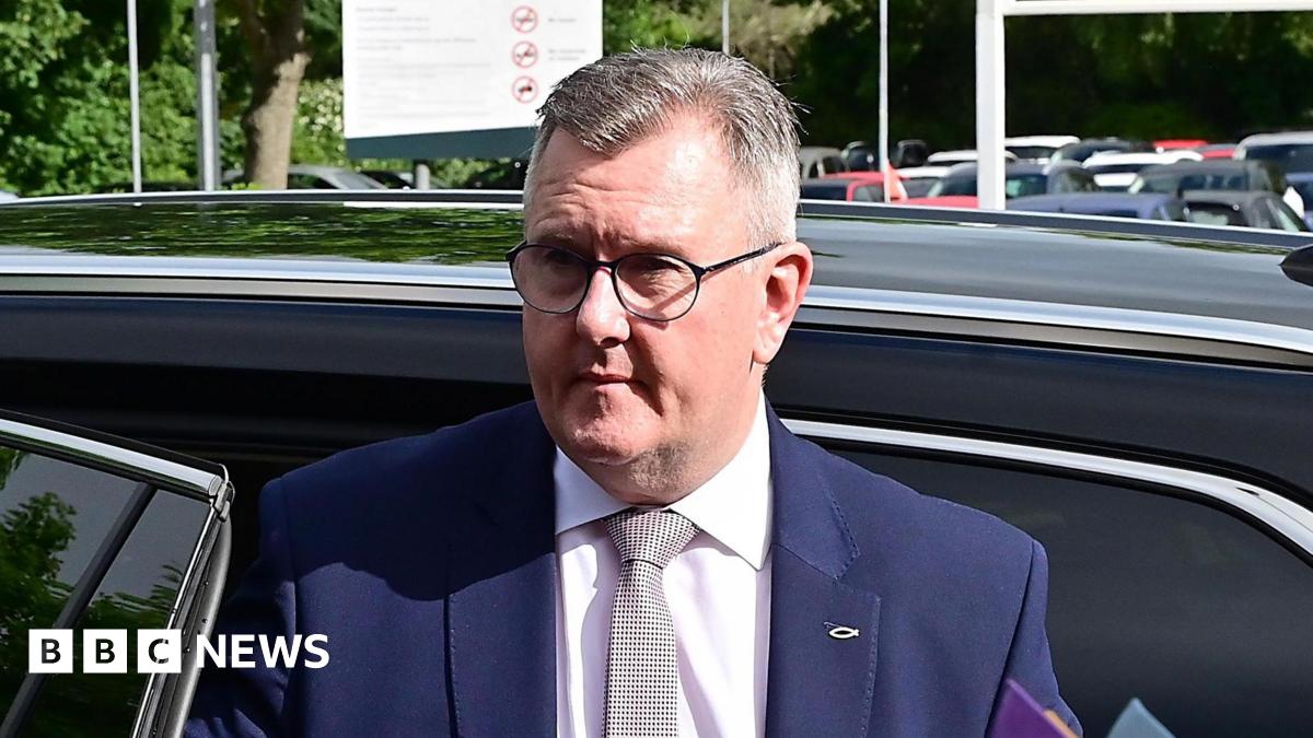 A man exiting a car - he is standing up just at the open doorway of the back seat. He has short greying hair and is wearing glasses. The top of his torso is visible, he's wearing a blue suit jacket, white shirt and tie with a black lined pattern on a white background. The car he is exiting is black. In the background the tops of other cars in a car park are visible