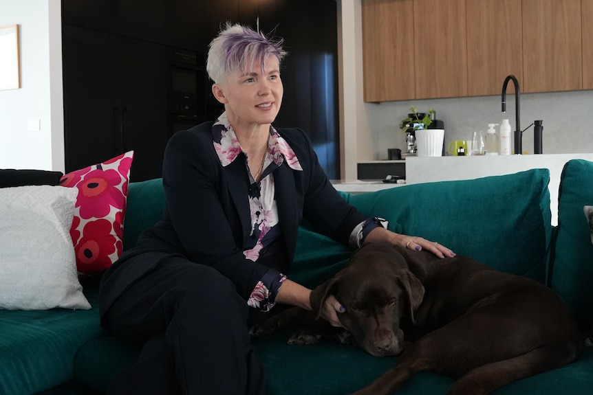 Sue with purple hair and a colourful patterned shirt sits on a couch, patting a large brown dog.
