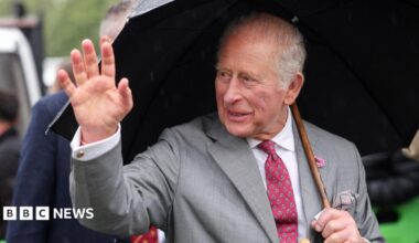 King Charles smiles and waves while sheltering under an umbrella