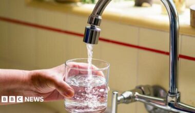 The image shows a close‑up of a person holding a clear glass under a kitchen faucet while fresh water flows into it. The hand is positioned securely around the glass, which is already partially filled. The faucet is metallic and curved, and the water stream appears clean and steady. In the background, the kitchen counter and tiled backsplash are slightly out of focus.