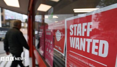 An image of a man walking past a red sign in a window saying staff wanted