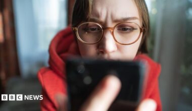 Woman wearing glasses looks at phone which is prominent in the foreground.