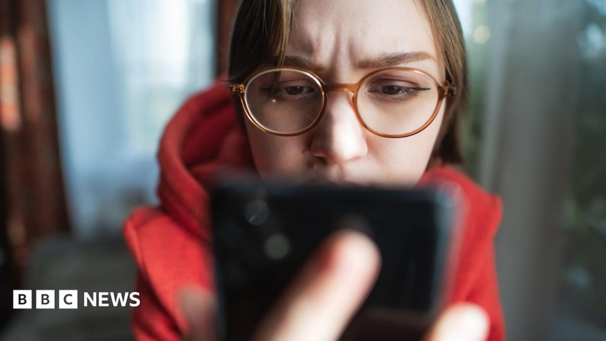 Woman wearing glasses looks at phone which is prominent in the foreground.
