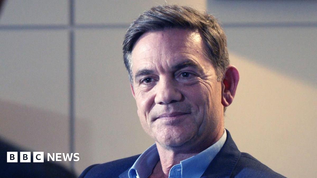 John Michie wearing a dark blue suit jacket and blue shirt. He has short dark hair.