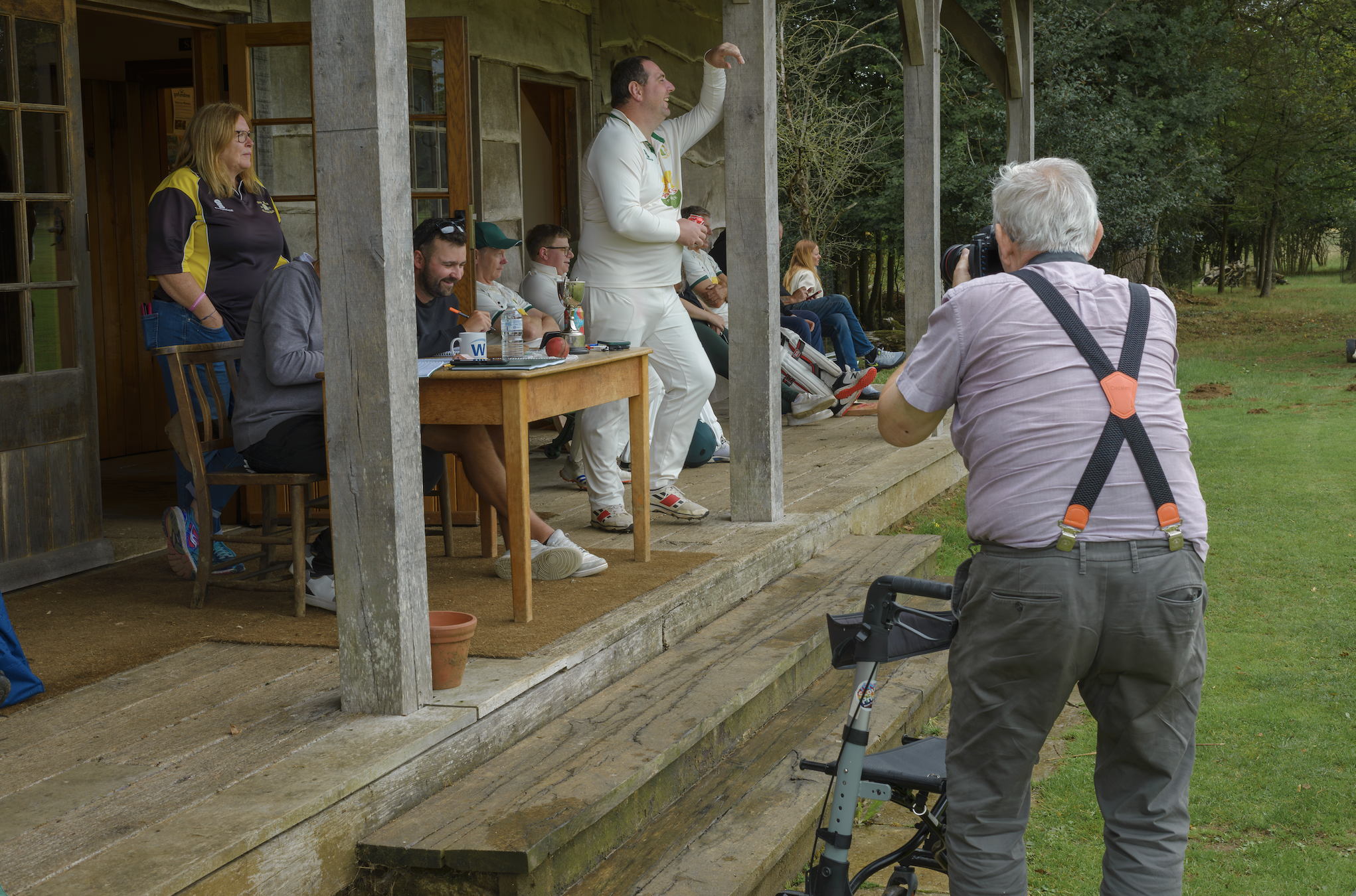 Martin Parr, wearing braces and using a walking frame, photographs flag bunting flapping over a group seated in camping chairs on a village green.