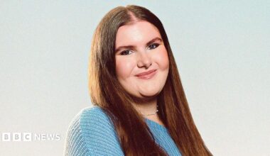A professional headshot of Katie Hudson who has long brown hair. She is wearing a silver necklace and blue jumper and is smiling.