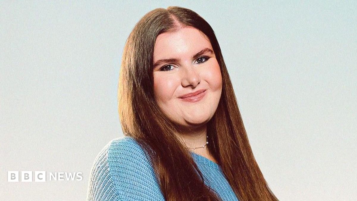 A professional headshot of Katie Hudson who has long brown hair. She is wearing a silver necklace and blue jumper and is smiling.