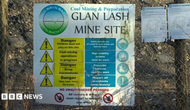 A sign at the entrance to Glan Lash opencast coal mine near Llandybie, Carmarthenshire. The white sign is attached to a metal link fence, with the words 'Glan Lash mine site'. The sign also has a yellow triangle caution sign and an image of a figure wearing a safety helmet.