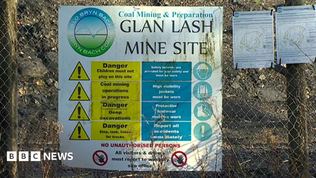 A sign at the entrance to Glan Lash opencast coal mine near Llandybie, Carmarthenshire. The white sign is attached to a metal link fence, with the words 'Glan Lash mine site'. The sign also has a yellow triangle caution sign and an image of a figure wearing a safety helmet.