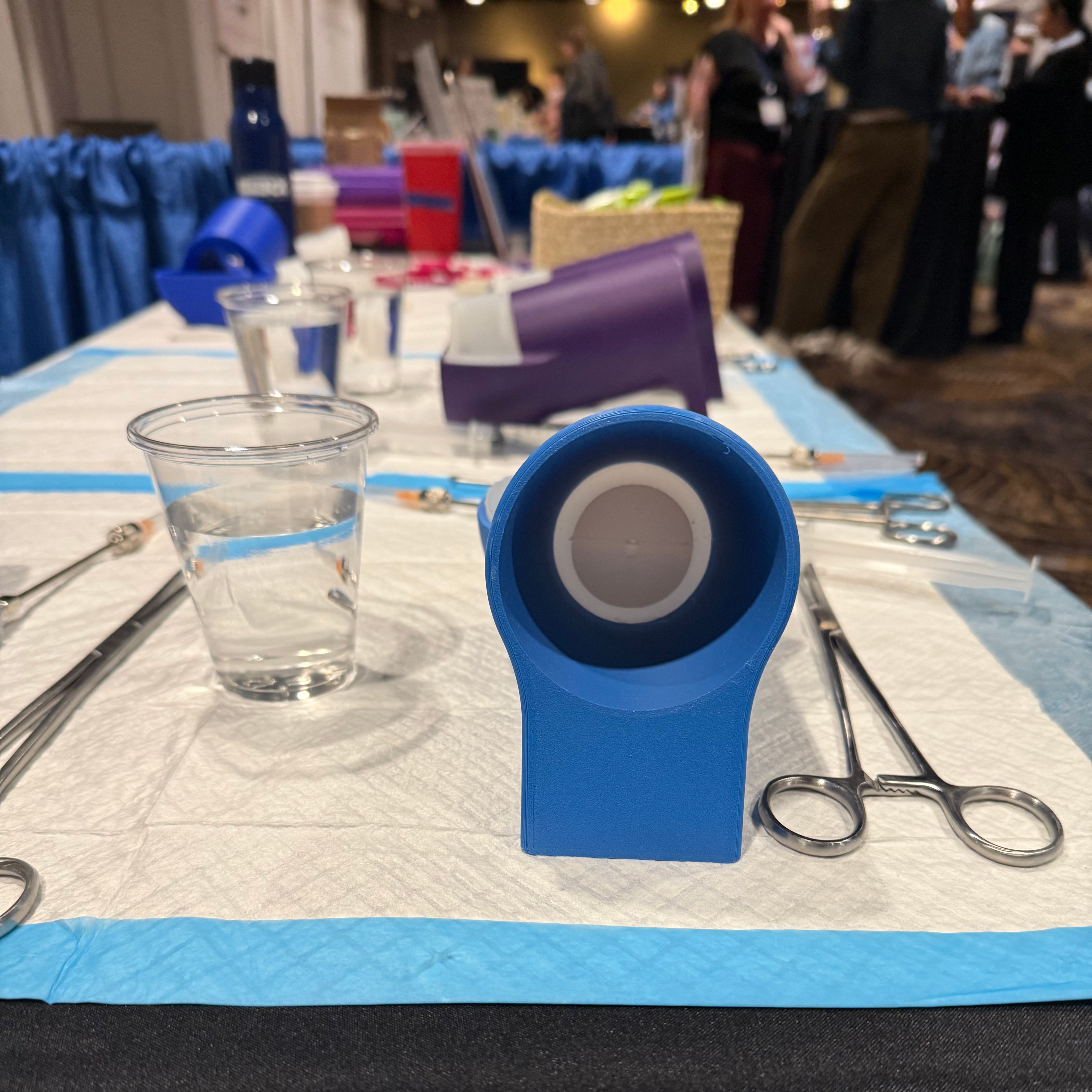 Side view of Kakaiya’s pelvic model, a blue plastic base holding a white silicone cervix, with forceps lying alongside.