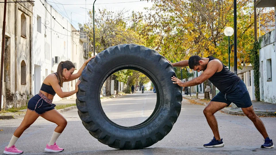 Two people in athletic wear push a large tire in the middle of a street lined with trees and buildings