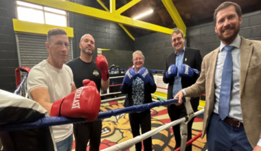 Three men and a woman standing in a boxing ring, and one man standing just outside it. The ones inside are wearing boxing gloves and they are all smiling.