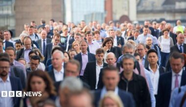A large, dense crowd of commuters, mostly dressed in business and smart-casual attire, walking towards the camera on a city street or bridge, with buildings visible in the blurred background.