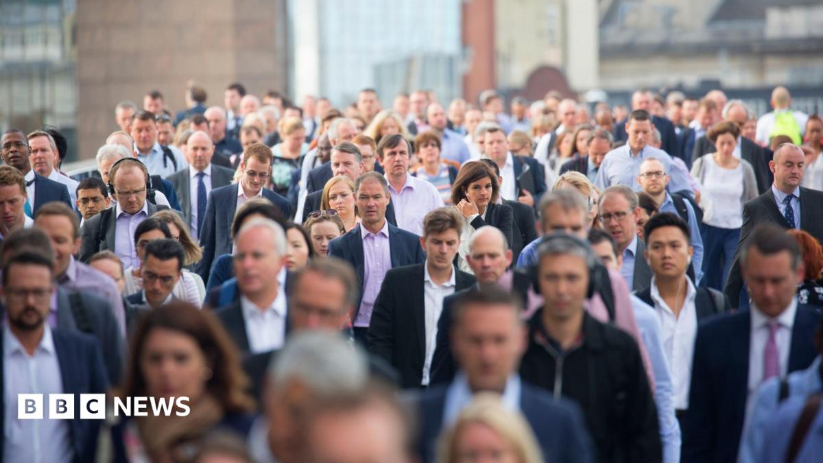 A large, dense crowd of commuters, mostly dressed in business and smart-casual attire, walking towards the camera on a city street or bridge, with buildings visible in the blurred background.
