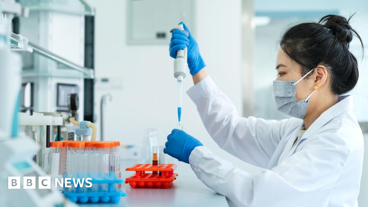 A laboratory setting with a person in a white lab coat working at a bench. The person is wearing protective gloves and a face mask while carefully using a pipette to transfer liquid into a small test tube. Several racks of test tubes with coloured liquids are arranged neatly on the work surface in the foreground. Scientific equipment and shelves are visible in the background, suggesting a clean and organised workspace