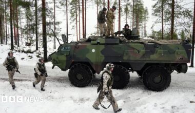 An armoured vehicle on a snow-covered road. There are three soldiers on top of the tank and three others walking along side it.
