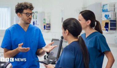 One male and two female nurses in blue scrubs stand in a hospital room.