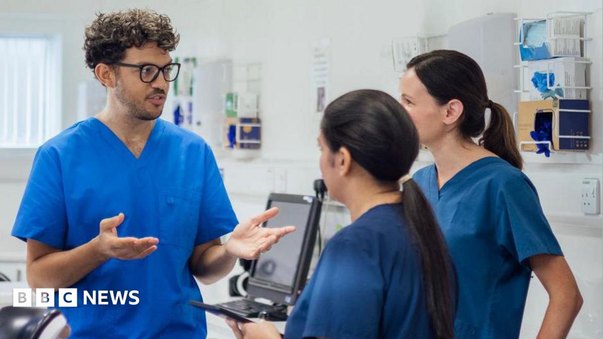 One male and two female nurses in blue scrubs stand in a hospital room.