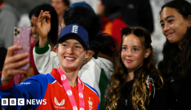 A mid-shot of Charlie standing in a crowd holding up a camera for a selfie and smiling along with two young female cricket fans