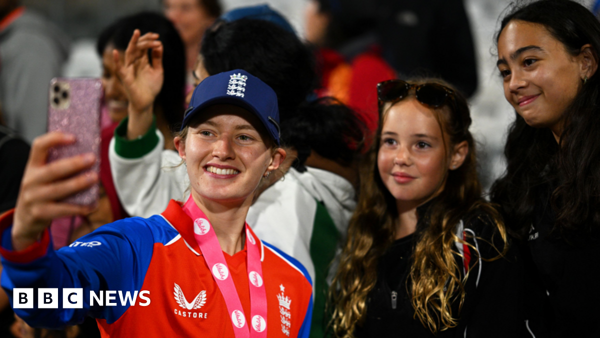 A mid-shot of Charlie standing in a crowd holding up a camera for a selfie and smiling along with two young female cricket fans