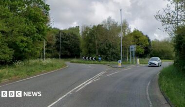 A google maps grab of a road, leading up to a roundabout. One silver car is visible. There is greenery on either side and in the centre of the roundabout.