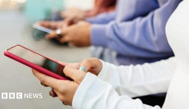 A close-up image of several young people using smartphones that appears to be indoor. The background is softly blurred, emphasising the hands and devices in the foreground.