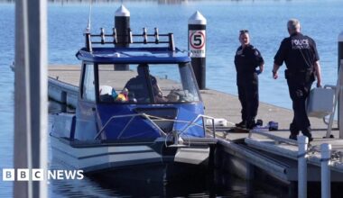 Two police officers stand on a jetty. One holds a large white box. Next to the jetty is parked a blue and white police boat, in which a third officer is sitting. All is surrounded by still and clear blue water