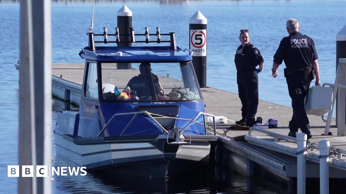Two police officers stand on a jetty. One holds a large white box. Next to the jetty is parked a blue and white police boat, in which a third officer is sitting. All is surrounded by still and clear blue water