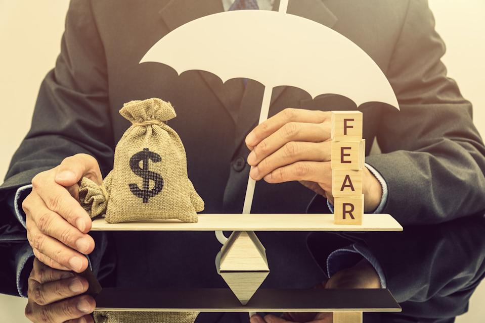 A money bag and wood blocks spelling "FEAR" on a scale with a person touching an umbrella covering the scale.