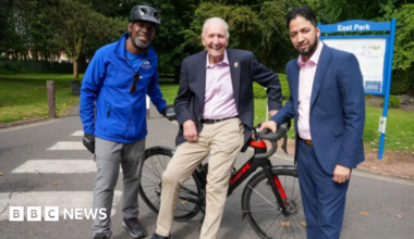 Three men, one seated on a stationary bike smile at the camera.