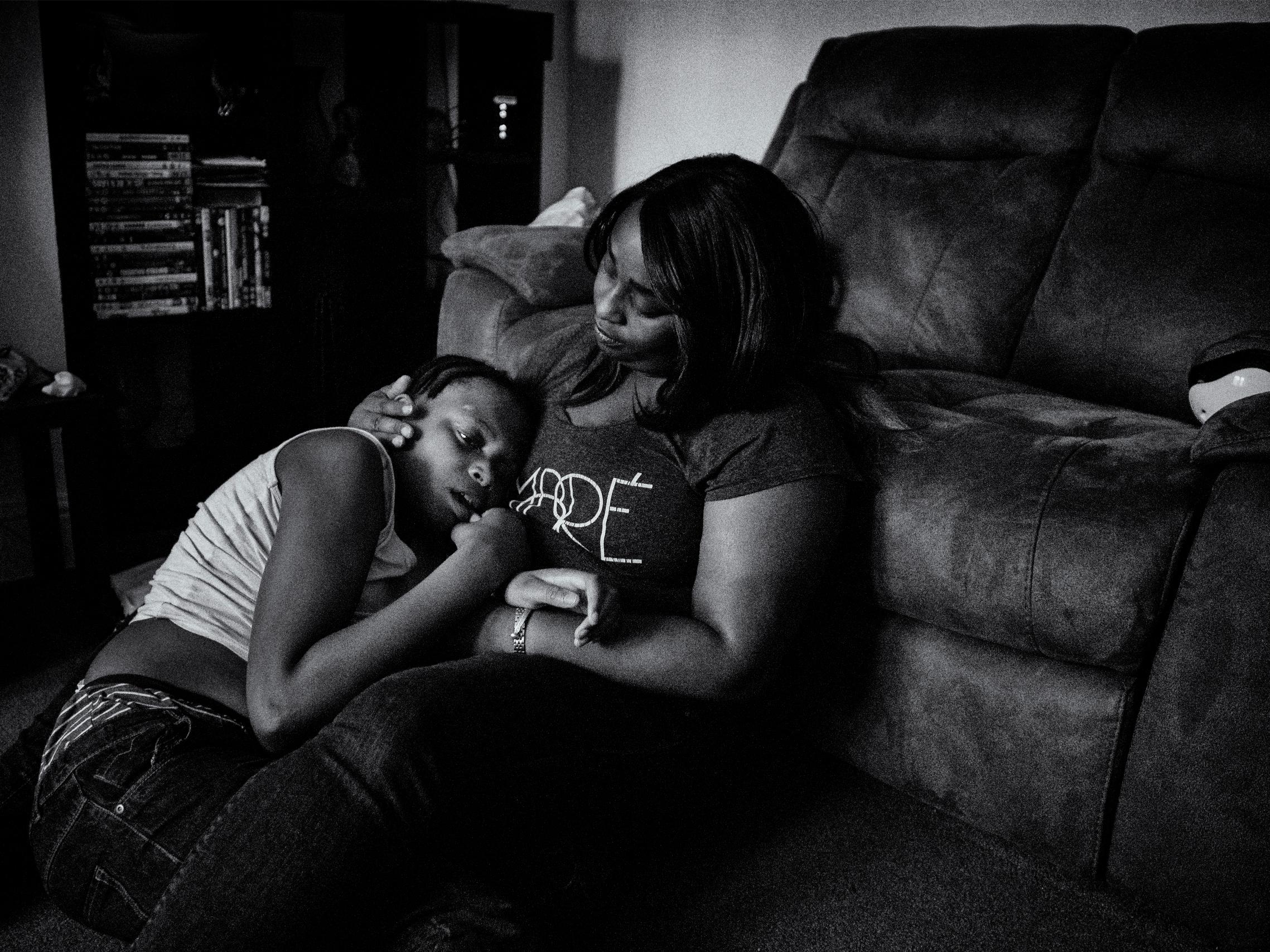A black and white photo of a woman sitting on the floor next to a couch, gently comforting a child who is leaning against her chest.