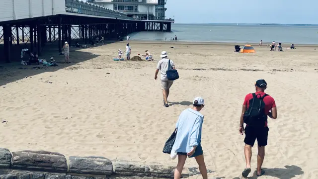 People are walking onto a beach front next to a pier on the left. An orange beach tent can be seen in the distance.