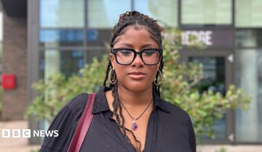 Mide Awosika wears glasses and a black shirt and is looking at the camera