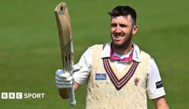 Craig Overton holds a bat towards the sky in his right arm, as he stands on the pitch during a match for Somerset