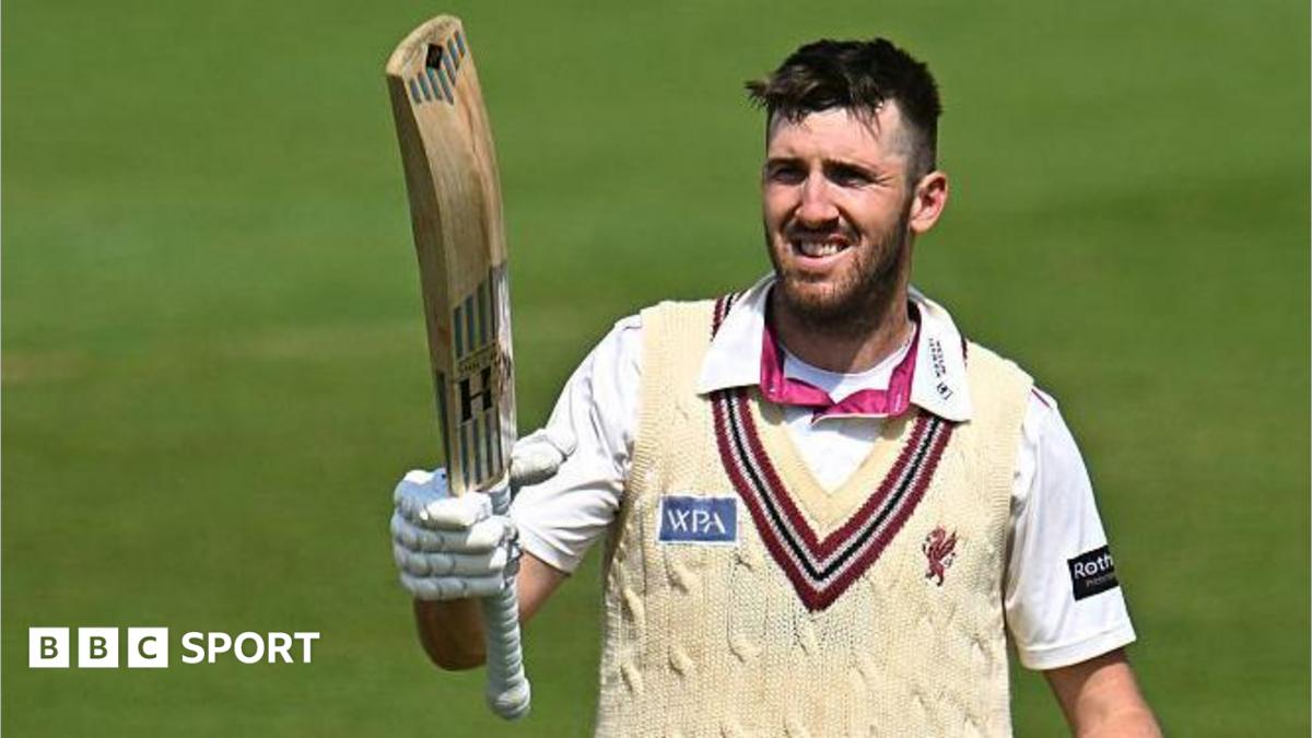 Craig Overton holds a bat towards the sky in his right arm, as he stands on the pitch during a match for Somerset