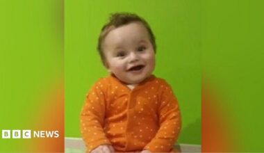 Undated image of the late baby Preston Davey. He has brown curly hair and brown eyes and is wearing an orange spotted baby grow. He is smiling.