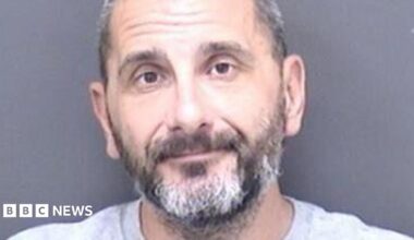 A custody shot of Matthew Ashley Foster-Smith. He has short hair and a beard and is wearing a grey jumper.