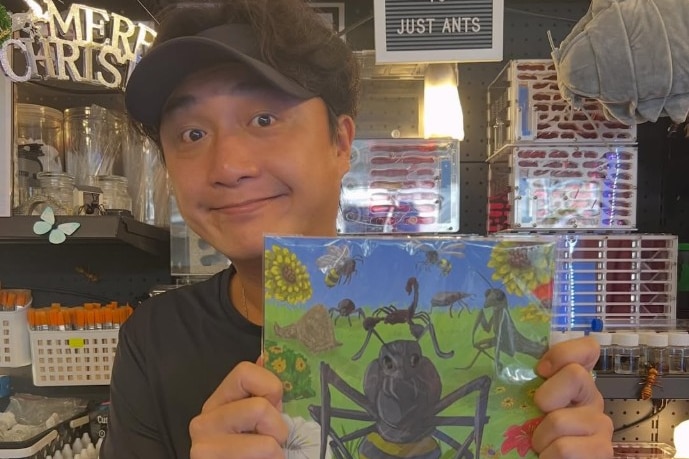 A man, standing in front of ant enclosures, holds up a book about ants.