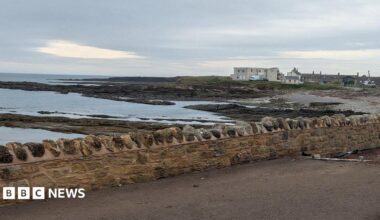 The coastal defence wall along Harbour Road in Beadnell, Northumberland. The stone wall is built near the shore. The sea can be seen behind it. A village can be seen in the distance.