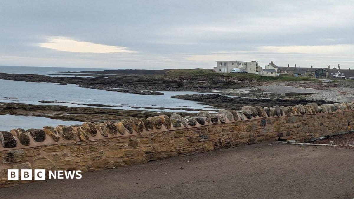 The coastal defence wall along Harbour Road in Beadnell, Northumberland. The stone wall is built near the shore. The sea can be seen behind it. A village can be seen in the distance.
