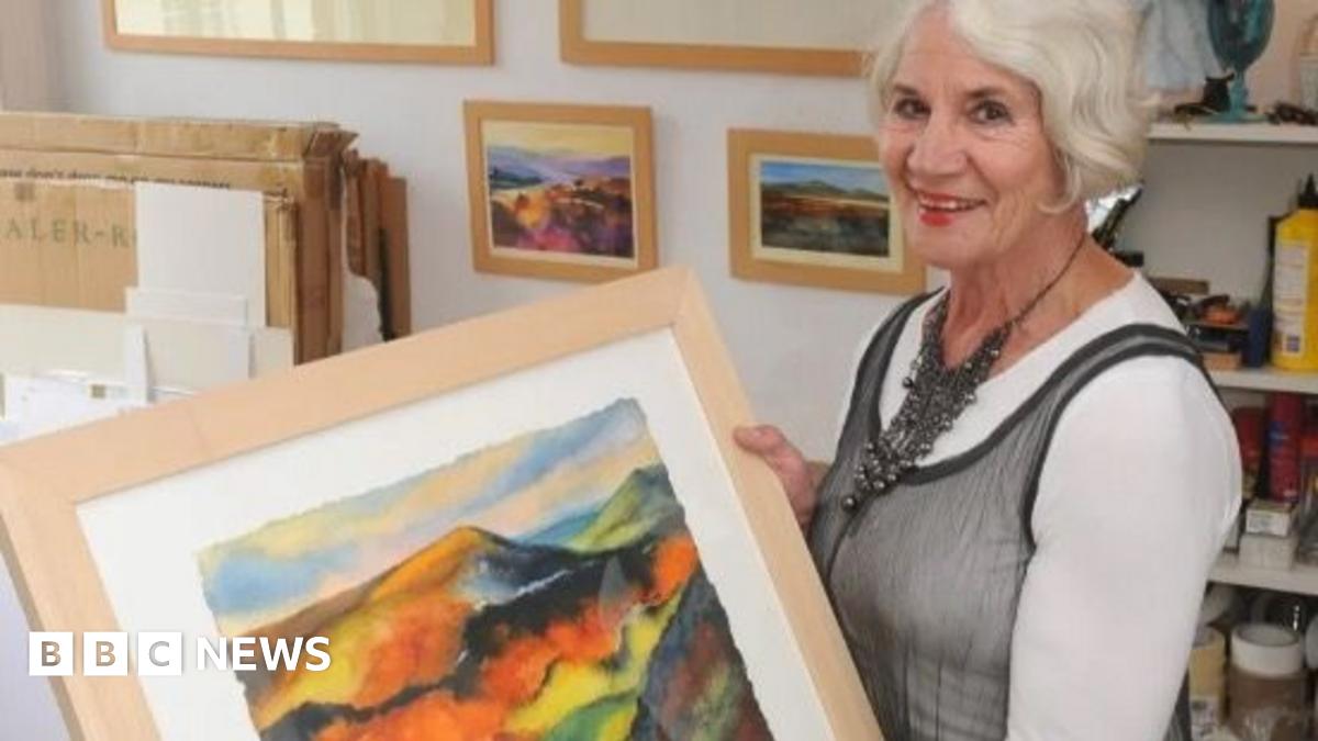 Woman with white hair holding a multi-coloured landscape painting showing hills and fields. She is wearing a grey dress with white top underneath