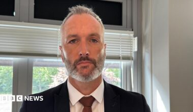 Det Con Lee Harshey-Jones. He looks at the camera with a neutral expression, and wears a white shirt, black suit jacket and bronze tie. He has short grey hair and short stubble.